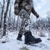 Person in winter camouflage gear wearing JHB06 heated boots walking through snow-covered forest